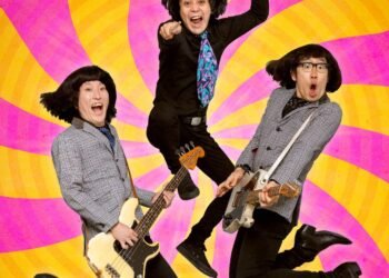 Photo of the three members of THE 50KAITENZ in mid-jump holding their instruments, with each person prominently displaying their individuality while maintaining a cohesive group aesthetic that hint at a 1960s inspiration. They are set against a vibrant background combining colours of yellowish/orange and pink.