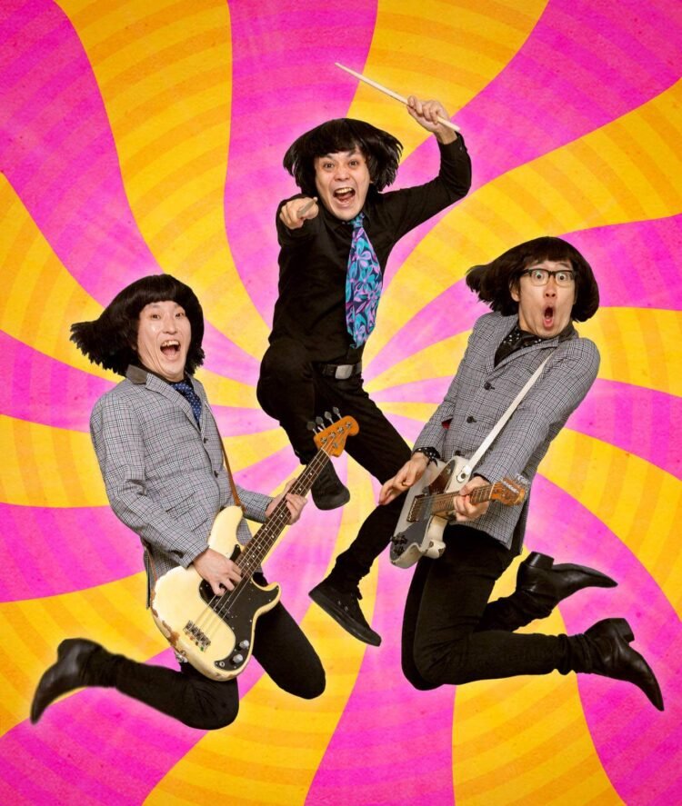 Photo of the three members of THE 50KAITENZ in mid-jump holding their instruments, with each person prominently displaying their individuality while maintaining a cohesive group aesthetic that hint at a 1960s inspiration. They are set against a vibrant background combining colours of yellowish/orange and pink.