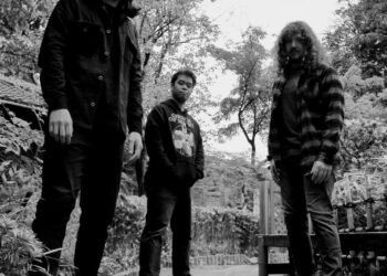 The photo is a black and white photograph of the three men of HEBI KATANA standing together outdoors, in what appears to be an overgrown garden. The background is filled with densely grown trees and bushes, and the ground is covered with irregularly shaped tiles.