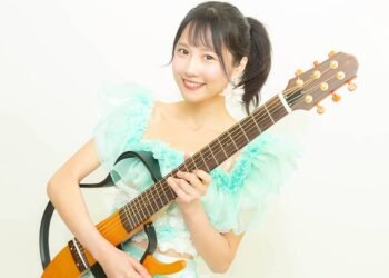 MION confidently holds her signature guitar. She is dressed in a light blue/greenish and white outfit and has her hair tied in a pony tail.