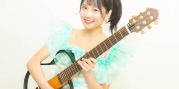 MION confidently holds her signature guitar. She is dressed in a light blue/greenish and white outfit and has her hair tied in a pony tail.