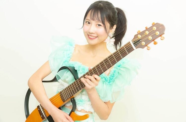 MION confidently holds her signature guitar. She is dressed in a light blue/greenish and white outfit and has her hair tied in a pony tail.