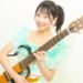 MION confidently holds her signature guitar. She is dressed in a light blue/greenish and white outfit and has her hair tied in a pony tail.