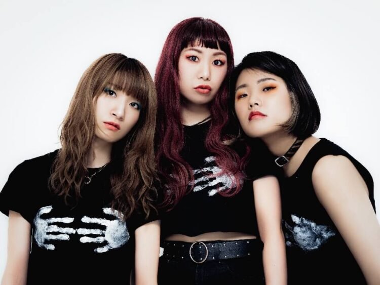 The photo features three women of THE LET'S GO's dressed in black T-shirts with white hand prints on the front. They are posed close together, looking directly at the camera.