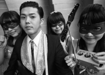 A black and white photo featuring the band Stompin’ Riffraffs, consisting of a man dressed in a suit and three masked ladies. They are standing near each other and carry their instruments. Stompin’ Riffraffs © Wild Records