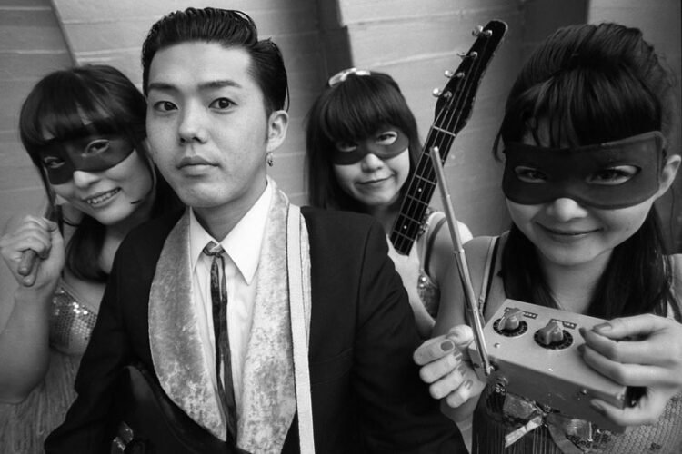 A black and white photo featuring the band Stompin’ Riffraffs, consisting of a man dressed in a suit and three masked ladies. They are standing near each other and carry their instruments. Stompin’ Riffraffs © Wild Records