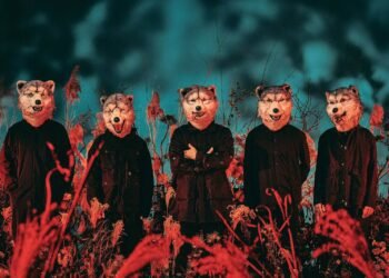 The photo depicts the five members of Man With A Mission wearing their signature wolf masks. They are dressed in black and standing in a line within a natural setting of what looks like tall, wild grass and plants. The photo consists of redish colours in the front and blue/green colours in the back.