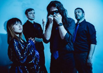 The photo depicts the four members of Re:O standing together against a blueish background. All band members are dressed predominantly in black, with three of them wearing purple ties and frontwoman Rio is dressed in what seems to be yukata or yukata-inspired garment in the colour dark blue/purple with white floral patterns. Photo by Agnieszka Rzesniowiecka.