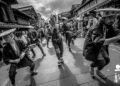 The photo is a black and white photograph displaying a performance of Seppuku Pistol in a street in Japan. The performers are dressed in traditional Japanese attire and holding instruments and dancing while there are audience standing around to watch the performance.