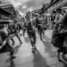 The photo is a black and white photograph displaying a performance of Seppuku Pistol in a street in Japan. The performers are dressed in traditional Japanese attire and holding instruments and dancing while there are audience standing around to watch the performance.