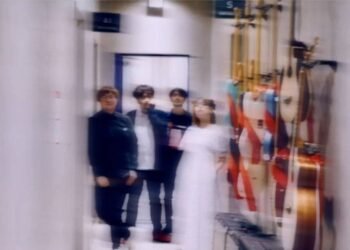 The four band members of Taffy are standing together in what appears to be a hallway, perhaps somwhere at a backstage venue. Most of the members are dressed in dark clothes, except Iris, who is wearing a white dress. Several musical instruments are mounted on the wall on the leftside.