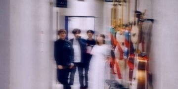 The four band members of Taffy are standing together in what appears to be a hallway, perhaps somwhere at a backstage venue. Most of the members are dressed in dark clothes, except Iris, who is wearing a white dress. Several musical instruments are mounted on the wall on the leftside.
