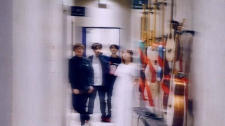 The four band members of Taffy are standing together in what appears to be a hallway, perhaps somwhere at a backstage venue. Most of the members are dressed in dark clothes, except Iris, who is wearing a white dress. Several musical instruments are mounted on the wall on the leftside.