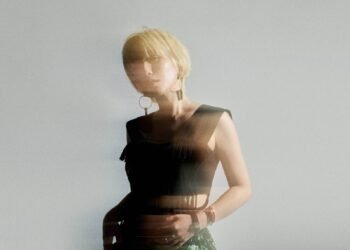The photo features Nao Yoshioka with blonde hair posing in a studio setting. She is wearing a black crop tank top and green trousers. There is an effect added to the photo giving an idea of movement and blurriness.