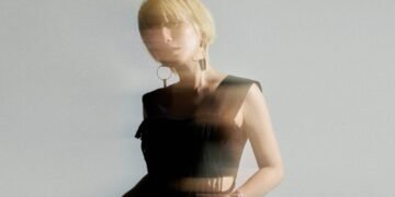 The photo features Nao Yoshioka with blonde hair posing in a studio setting. She is wearing a black crop tank top and green trousers. There is an effect added to the photo giving an idea of movement and blurriness.