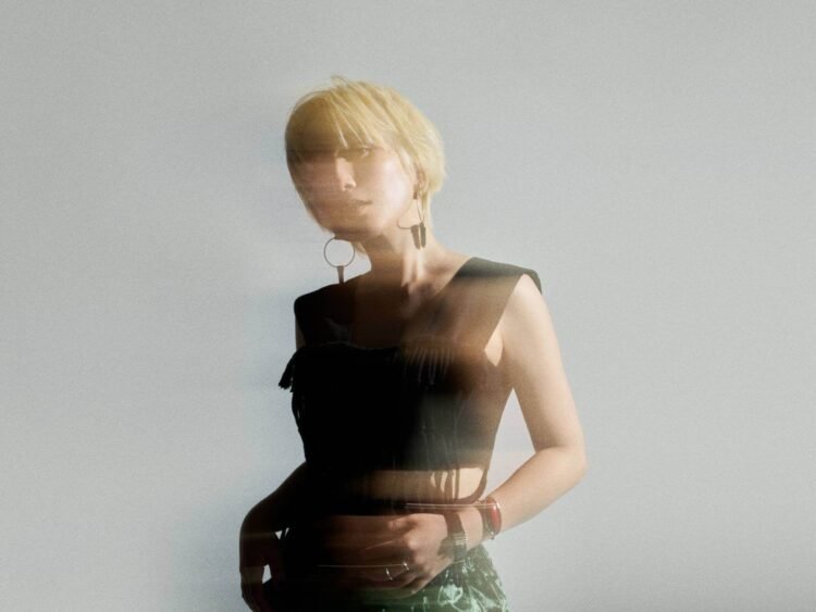 The photo features Nao Yoshioka with blonde hair posing in a studio setting. She is wearing a black crop tank top and green trousers. There is an effect added to the photo giving an idea of movement and blurriness.