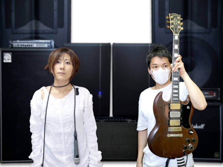 The photo features the two members of MELT-BANANA standing against a background of amplifiers and a white wall. Both are dressed in white outfits. The vocalist wears the microphone and cable around her neck as a necklace, while the guitarist wears a white mask and is holding his guitar up.