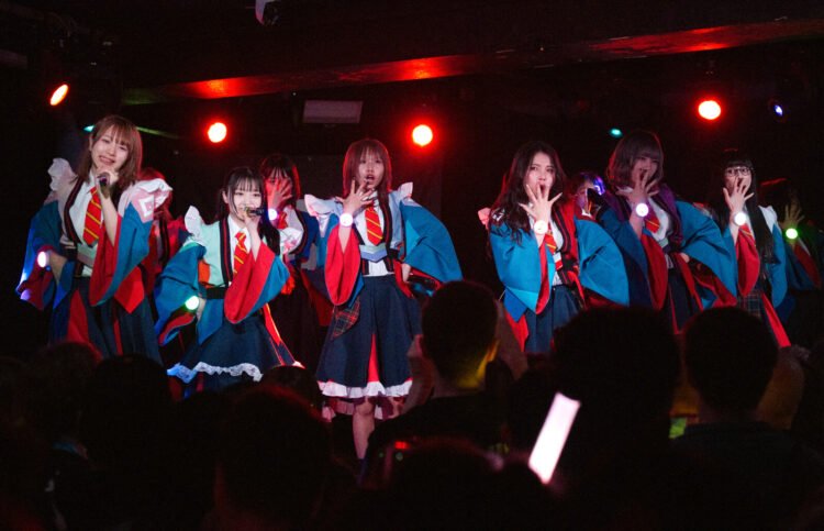 The photo pictures the group GANG PARADE dressed in red and blue uniforms engaged in a live performance on stage. Most of the performers are have their open hand in front of their mouth as a pose, while a couple are singing into a microphone. The performers are arranged in a row and the audience can be seen in the front in the shadow.