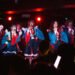 The photo pictures the group GANG PARADE dressed in red and blue uniforms engaged in a live performance on stage. Most of the performers are have their open hand in front of their mouth as a pose, while a couple are singing into a microphone. The performers are arranged in a row and the audience can be seen in the front in the shadow.