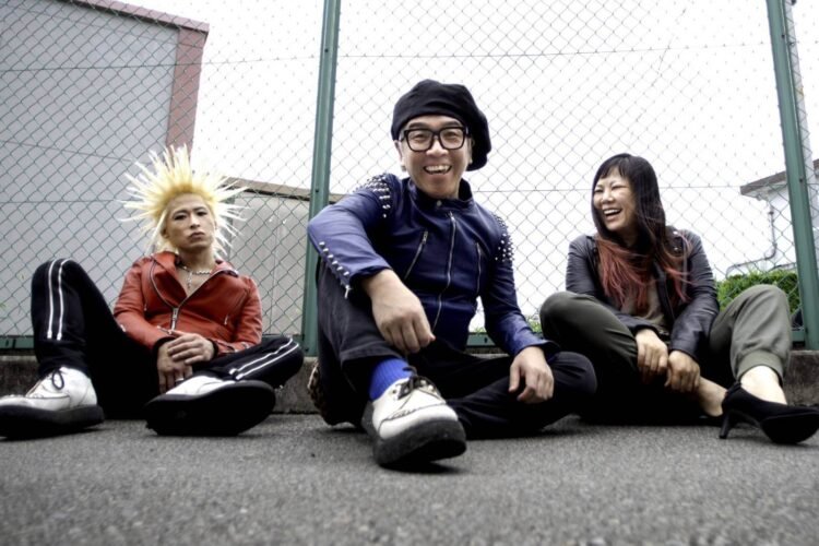 The photo shows the three band members of GNU MAD RUSSELL$ sitting on the ground in front of a fence. They are wearing leather long-sleeved jackets, all a different colours (bright red, blue and black). Two of the musicians are laughing, while the person with spiked blonde hair is looking seriously into the camera.