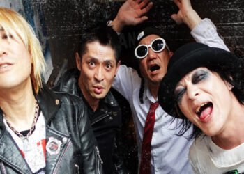 The four members of Japanese punk rock band FUNGUS are standing near each other before or after a concert. One wears a hat and thick black eye makeup and smiles, another one wears sun glasses and holds his arms up and opened his mouth, the other two have black leather jackets on and look seriously into the camera.