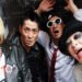 The four members of Japanese punk rock band FUNGUS are standing near each other before or after a concert. One wears a hat and thick black eye makeup and smiles, another one wears sun glasses and holds his arms up and opened his mouth, the other two have black leather jackets on and look seriously into the camera.