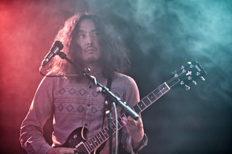 Kyotaro Miula of Minami Deutsch playing the guitar in front of the microfoon. The background, with green and redish colours with smoke, suggests a dreamy atmosphere.