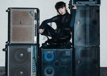 Yuko Araki is seated on a pile of large black speakers in a room with a white wall and a black floor. She is dressed entirely in dark clothing. Photo by Adì Putra