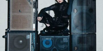 Yuko Araki is seated on a pile of large black speakers in a room with a white wall and a black floor. She is dressed entirely in dark clothing. Photo by Adì Putra