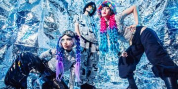 The three members of music project BIJŪ are actively posing against a backdrop which gives off a reflective, metallic look, similar to aluminum foil. The members are all dressed in a unique outfit, all wearing the same kind of shirt, that seems to be a newspaper design, the two ladies have the outfit complimented with colourful hair (pieces), a hat or goggles.