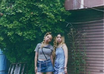 The photos features the two women of HYPER GAL standing side by side near a closed brown shutter that is covered with green plants. The photo is taken by Kensei Kataoka.