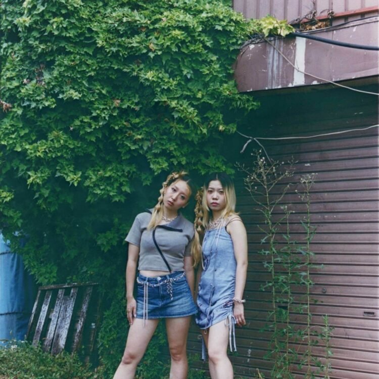 The photos features the two women of HYPER GAL standing side by side near a closed brown shutter that is covered with green plants. The photo is taken by Kensei Kataoka.