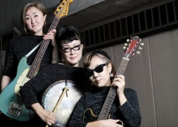 The photo features a group photo of three women of the garage rock band The 5.6.7.8’s, each holding a different musical instrument and wearing the same sparkly black outfits. © The 5.6.7.8's