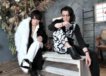 Mikaru and Yudai of G.L.A.M.S are sitting on a wooden bench, which is positioned against a backdrop of a gray wall dried plants. Both musicians are dressed in black and white clothing.