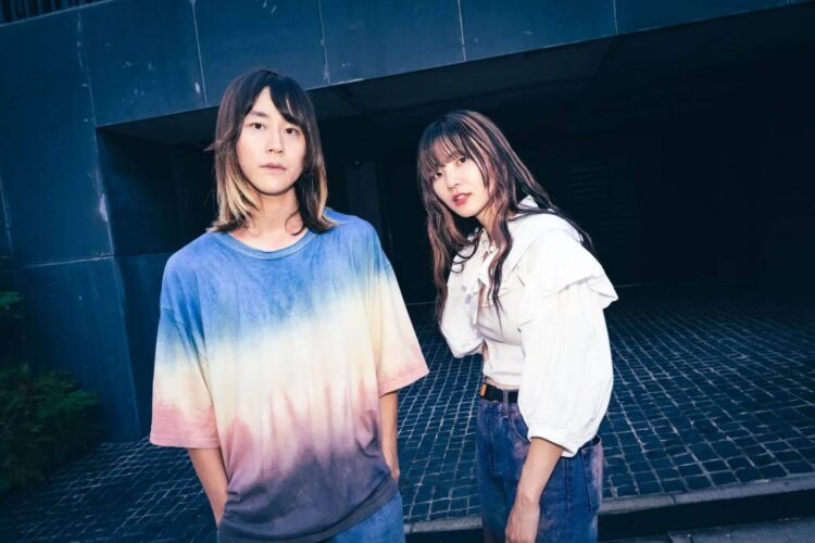 Band photo of SpecialThanks with Takaaki Yoshida and Misaki Anita standing on a place with stone tiles. Takaaki is wearing a tie-dye shirt and Misaki is wearing a white blouse and jeans.