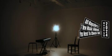Still from 烏兎 -uto-'s music video for the song "声 (Koe)", which is directed by Yusuke Arai (PLN2). The still is the end scene of the music video of a dimly lit room with a piano keyboard, chairs and a single spotlight as the musicians left the room after performing their song.