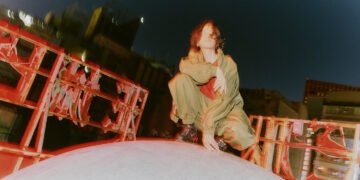 The photo shows the musician Kenshi Yonezu posing on top of a large white round object surrounded by red structures of neon signs, it seems to be located on top of a building. There are other buildings in the background.