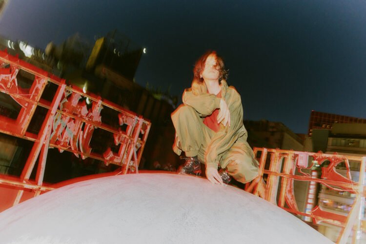 The photo shows the musician Kenshi Yonezu posing on top of a large white round object surrounded by red structures of neon signs, it seems to be located on top of a building. There are other buildings in the background. © Kenshi Yonezu | Photography by Kento Mori