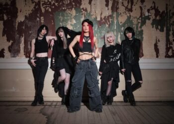 The photo shows the five members of BRIDEAR posing in what seems an old building with ripped off wallpaper and a wooden floor that has water damage. They are all dressed in black attire, although each member have their own unique look. In front stands the vocalist Kimi.