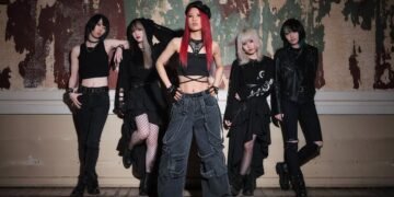 The photo shows the five members of BRIDEAR posing in what seems an old building with ripped off wallpaper and a wooden floor that has water damage. They are all dressed in black attire, although each member have their own unique look. In front stands the vocalist Kimi.