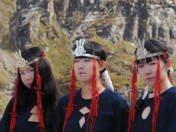 Photo of the tree ladies of KUUNATIC standing side by side wearing black dressed and silver headpieces and red cords that seem to be an artistic and symbolic interpretation, maybe inspired by sjamanistic or tribal styles. Photography by Celine Fougerouse