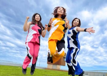 The three members of Shone Knife are jumping in the air on a small grassy field while smiling, the photo in this case is cropped. All members are dressed in an outfit of the same sort of design with white and black but each have a different third colour, pink, yellow and blue.