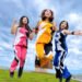The three members of Shone Knife are jumping in the air on a small grassy field while smiling, the photo in this case is cropped. All members are dressed in an outfit of the same sort of design with white and black but each have a different third colour, pink, yellow and blue.