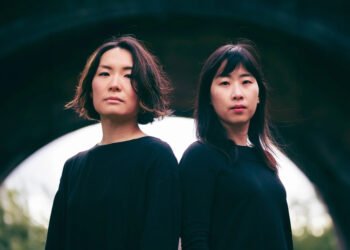 Photo of the Japanese performance artist Masumi Saito and Taiwanese musical alchemist An-Ting standing next to each other. They are dressed identically in a black sweater and stand in front of a small tunnel, bridge or stone door arch.
