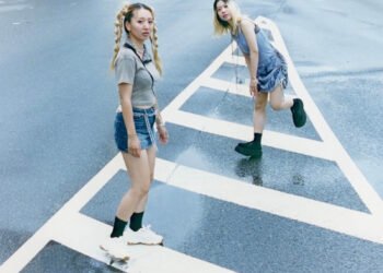 The photo features the two women of HYPER GAL standing in the middle of the street on white road markers as a white car just passes them. The photo is taken by Kensei Kataoka.