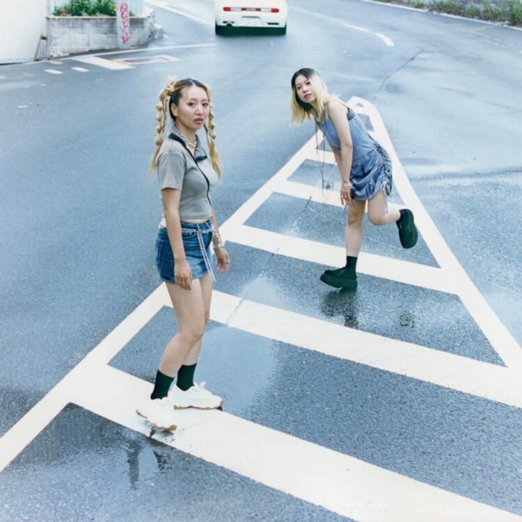 The photo features the two women of HYPER GAL standing in the middle of the street on white road markers as a white car just passes them. The photo is taken by Kensei Kataoka.