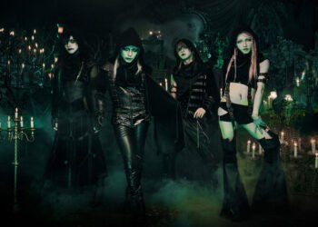 The photo features the four members of JILUKA, dressed in distinct dark, gothic attire. They are posing in a dark setting with chandeliers and numerous lit candles creating an eerie ambiance.