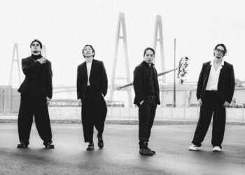 A black-and-white photo of the band Survive Said The Prophet standing on a road near a bridge. The bandmembers are all dressed in dark/black suits, posing all in a different way but facing the camera.