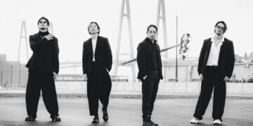 A black-and-white photo of the band Survive Said The Prophet standing on a road near a bridge. The bandmembers are all dressed in dark/black suits, posing all in a different way but facing the camera.