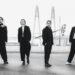 A black-and-white photo of the band Survive Said The Prophet standing on a road near a bridge. The bandmembers are all dressed in dark/black suits, posing all in a different way but facing the camera.